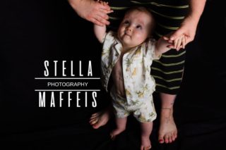 Standing baby looking up at his mother while holding her hands.