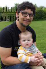 A dark curly haired man in black glasses, smiling at the camera while sitting outside on the grass, holding a chubby cheeked baby.