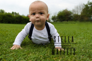 A baby sitting on the grass outside, with a funny frown and suspenders.