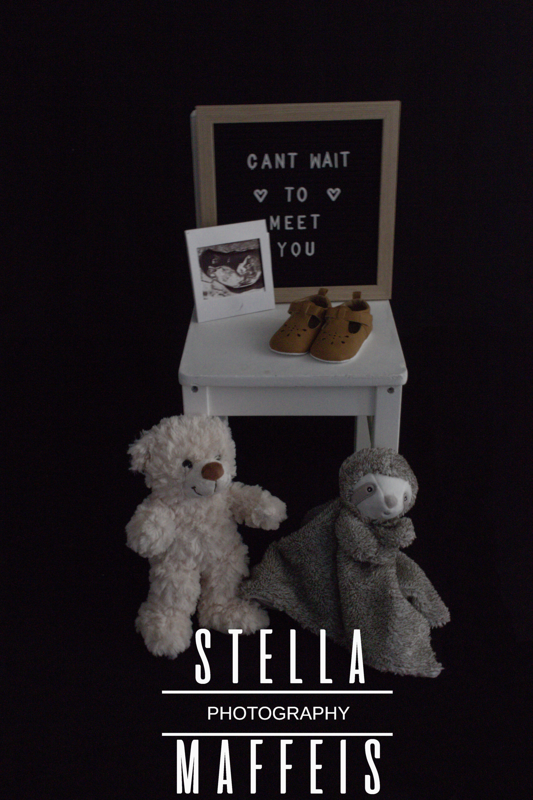 Photography shoot props, a little white chair with two teddys, a pair of little baby shoes, a sonogram image and a sign which reads "Can't wait to meet you".