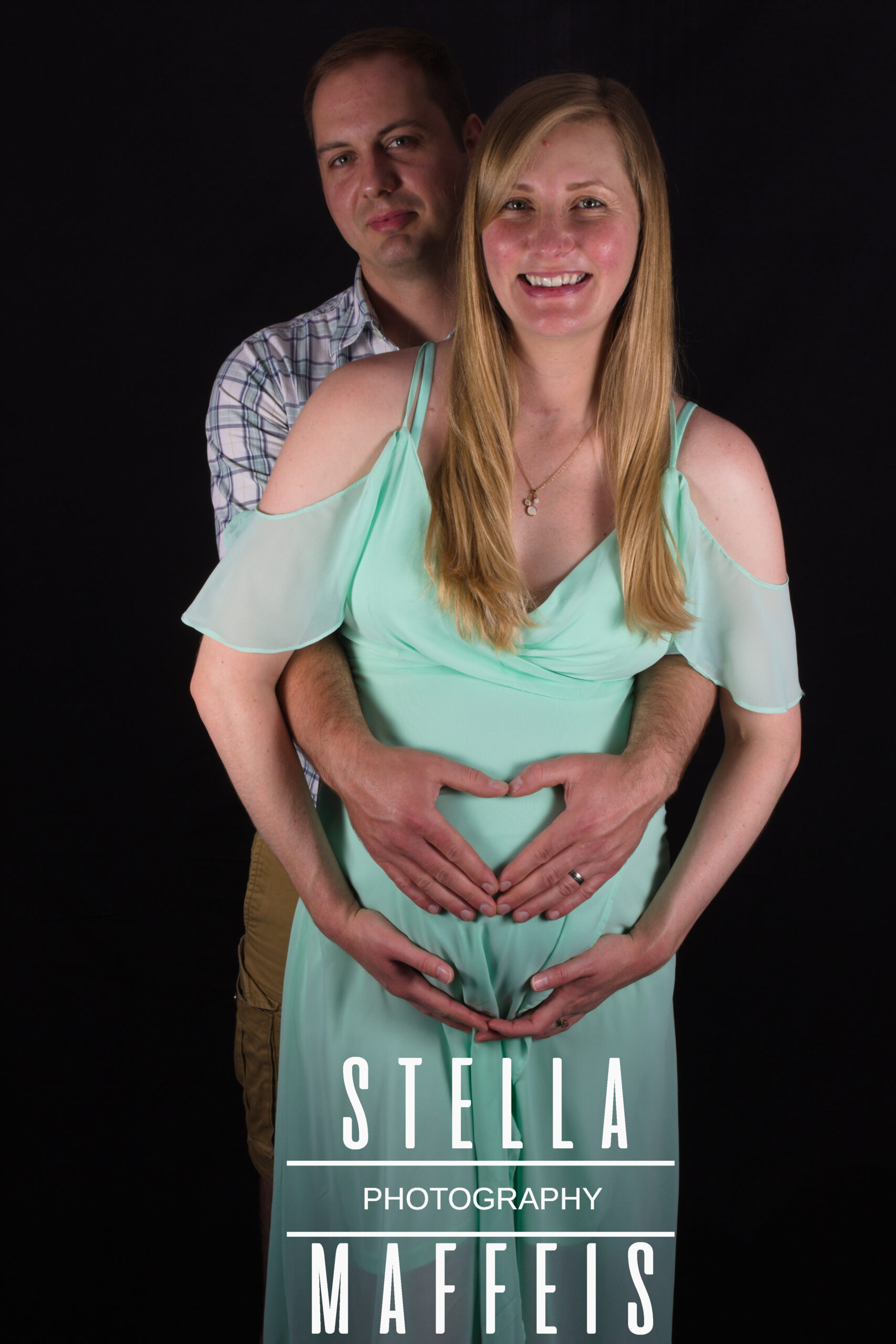 A pregnant woman holding her baby bump and her husband standing behind her, making a heart shape on her belly.