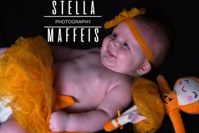 A smiling baby wearing an orange tutu and pumpkin headband with a halloween dressed doll.